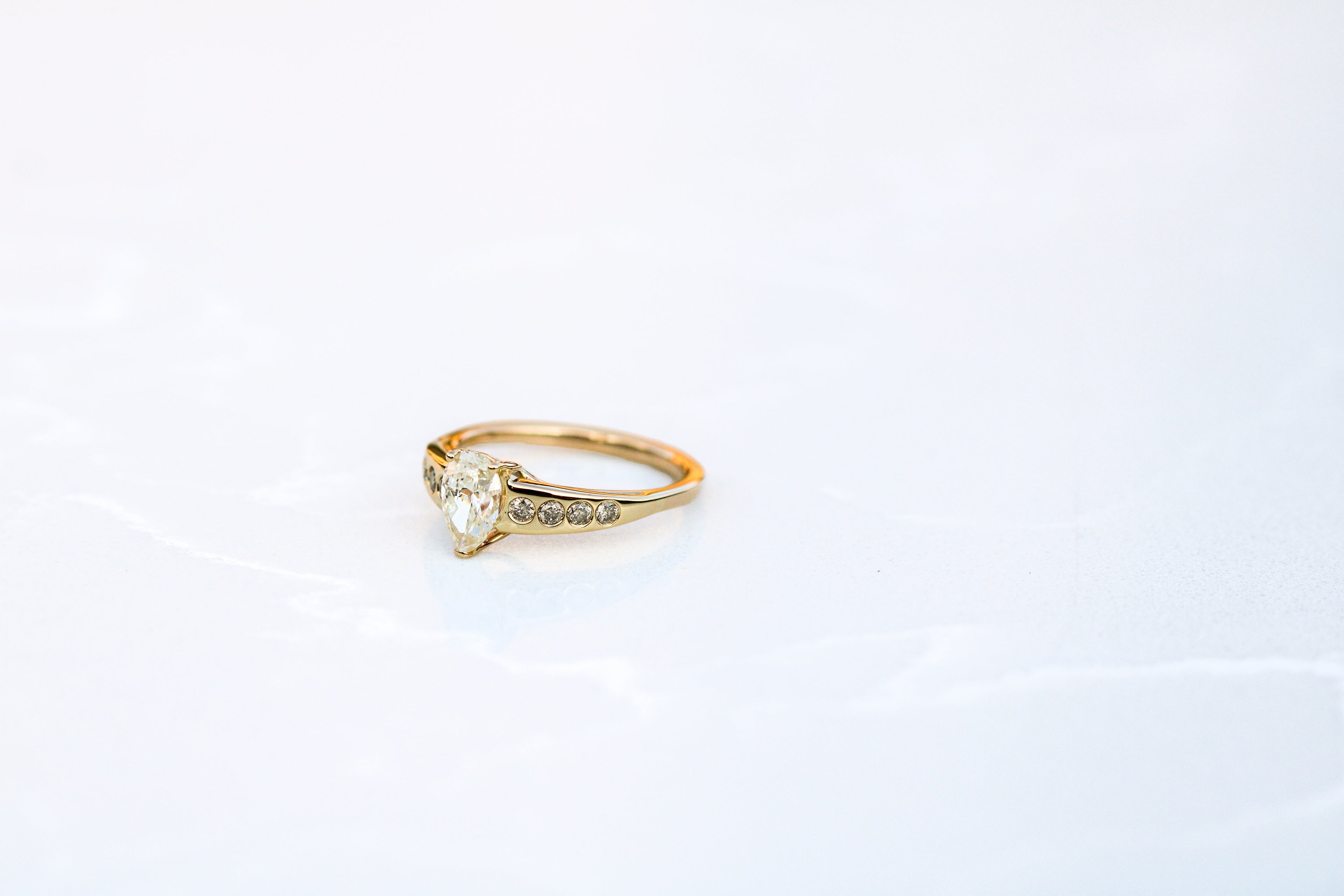 yellow gold ring with prong set pear shaped diamond center stone and 4 flush set round diamonds on each side.